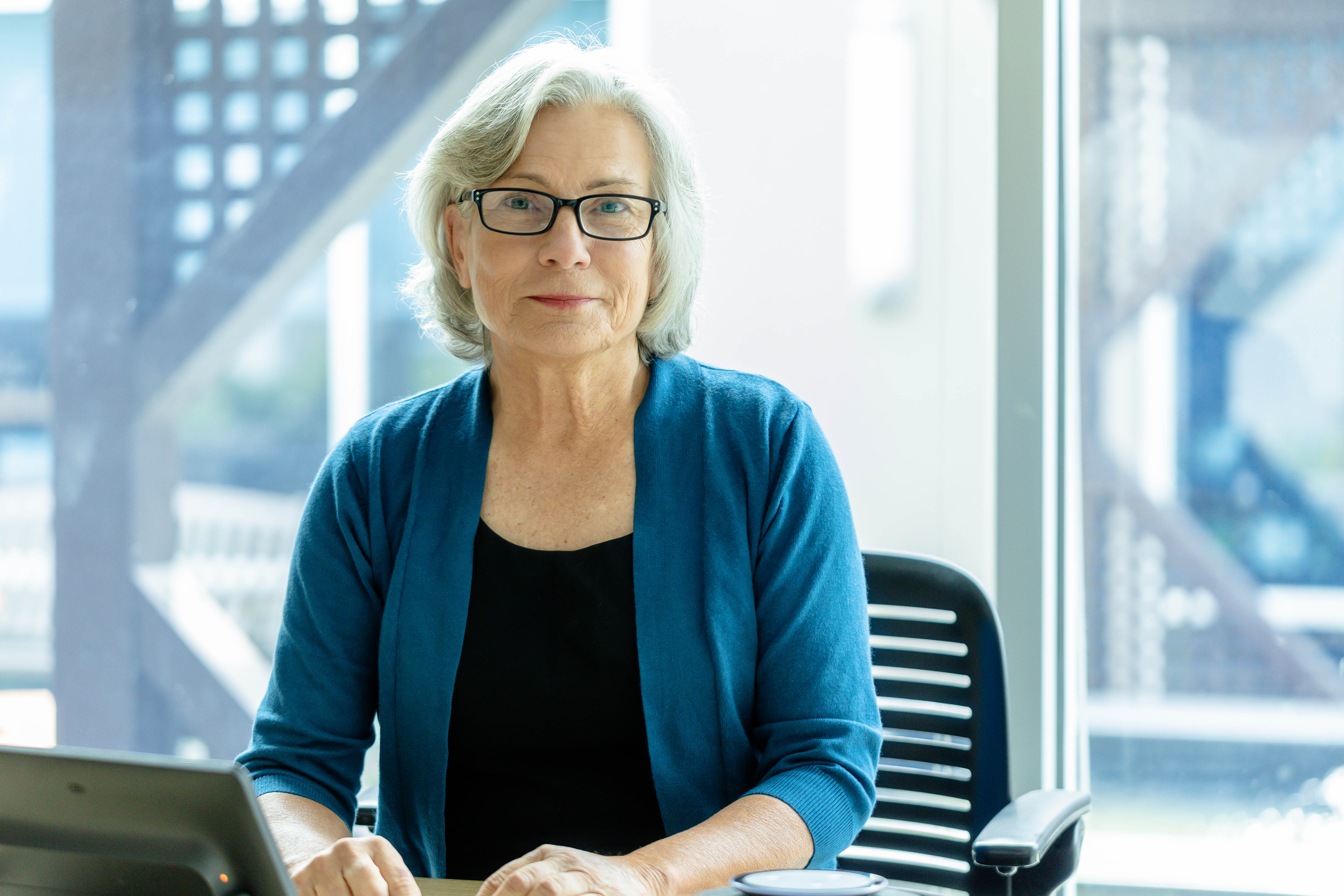 Person with grey hair, wearing glasses and a blue cardigan, seated at a desk.