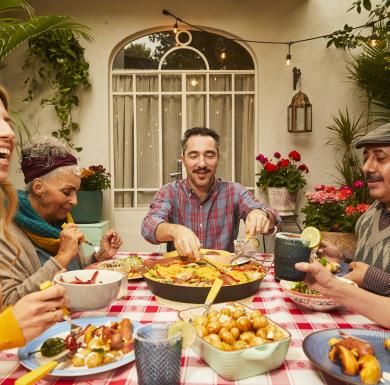 A group gathered outdoors sharing a meal with dishes like paella, potatoes, and vegetables.