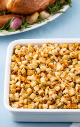 A casserole dish full of stuffing with a roasted turkey in the background, sitting on a blue clothed table.