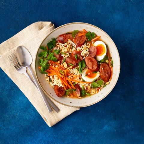 Bowl of ramen with spicy turkey sausage, eggs, carrots, cilantro, and peanuts on a blue background.