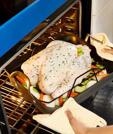 Turkey in a roasting pan with veggies being placed in a blue oven.