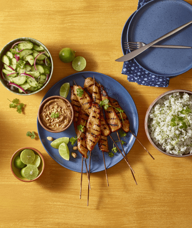 A meal with grilled turkey satays, rice, cucumber salad, peanut sauce and lime on a wooden table.