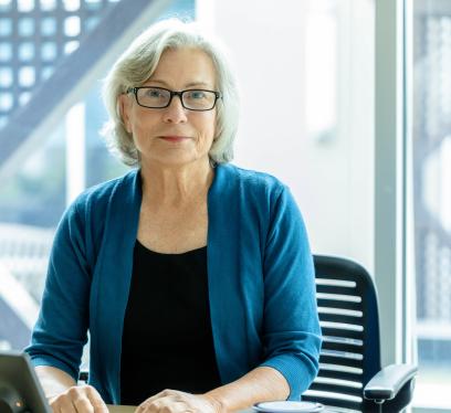 Person with grey hair, wearing glasses and a blue cardigan, seated at a desk.