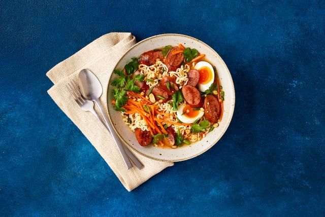 Bowl of ramen with spicy turkey sausage, eggs, carrots, cilantro, and peanuts on a blue background.