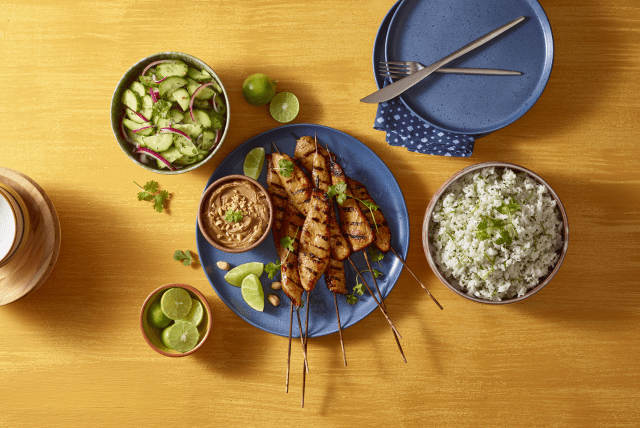 A meal with grilled turkey satays, rice, cucumber salad, peanut sauce and lime on a wooden table.